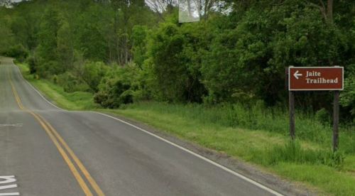 A road with a sign pointing left to "Jaite Trailhead," surrounded by lush green trees and grass.