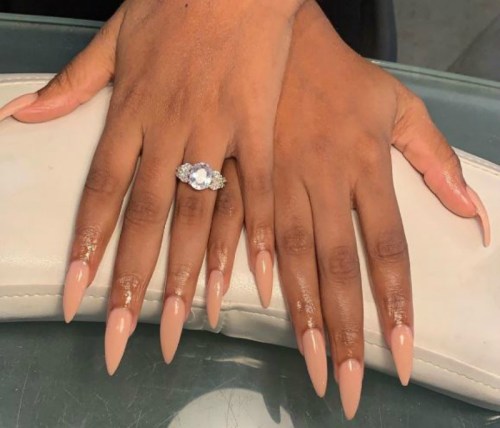 Close-up of two hands with long, pointed nude nails and a sparkling ring on one finger, resting on a white surface.