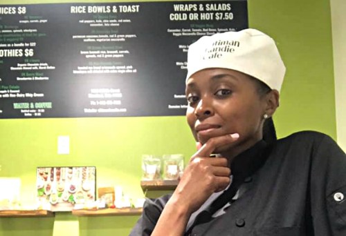 A chef in a white hat poses thoughtfully in a cafe with a menu displayed on the wall behind her.