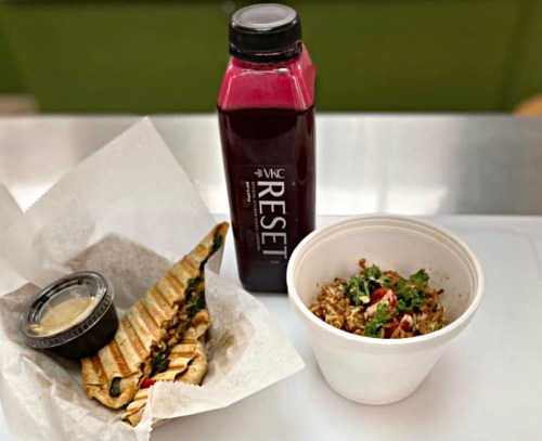 A grilled wrap with greens, a small container of sauce, a bowl of grain salad, and a bottle of juice.