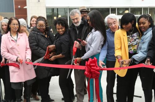 A diverse group of people gathers to cut a red ribbon at a ceremony, celebrating a new opening or event.