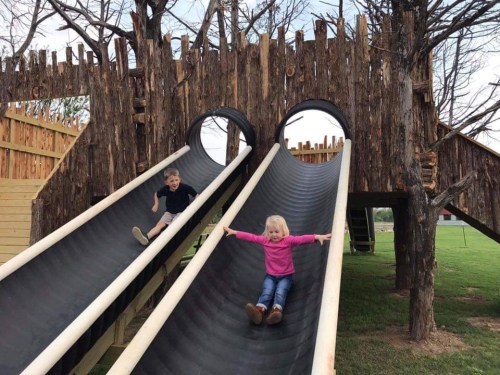 Two children slide down a wooden playground slide, surrounded by a rustic, tree-like structure.