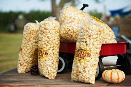 Bags of kettle corn stacked beside a red wagon, with a pumpkin and pinecones on a wooden table outdoors.