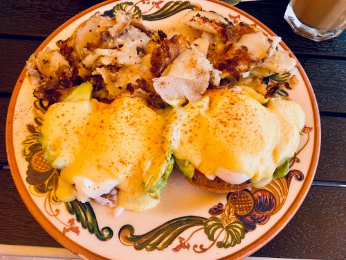 A plate of eggs Benedict with avocado and seasoned hash browns, served on a decorative dish.