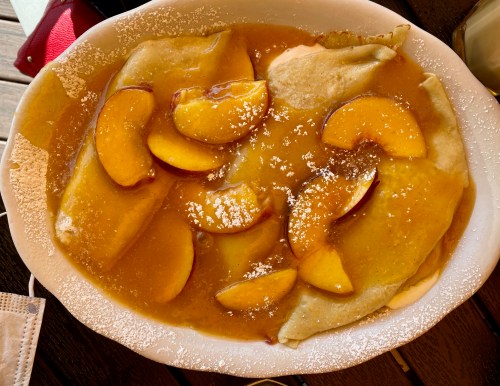 A plate of crepes topped with sliced peaches and a sweet sauce, dusted with powdered sugar.