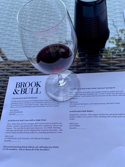 A glass of red wine sits on a table next to a menu featuring various wines from Brook & Bull.