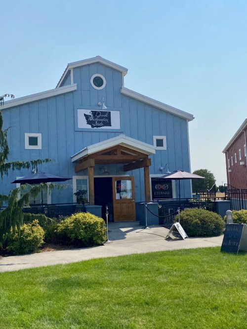 A blue barn-style building with a wooden entrance, surrounded by greenery and outdoor seating.