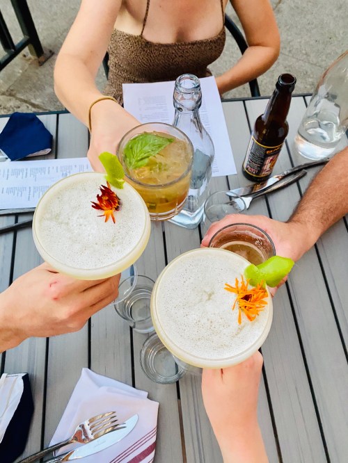 Three cocktails with garnishes are raised in a toast, surrounded by hands and a table setting.