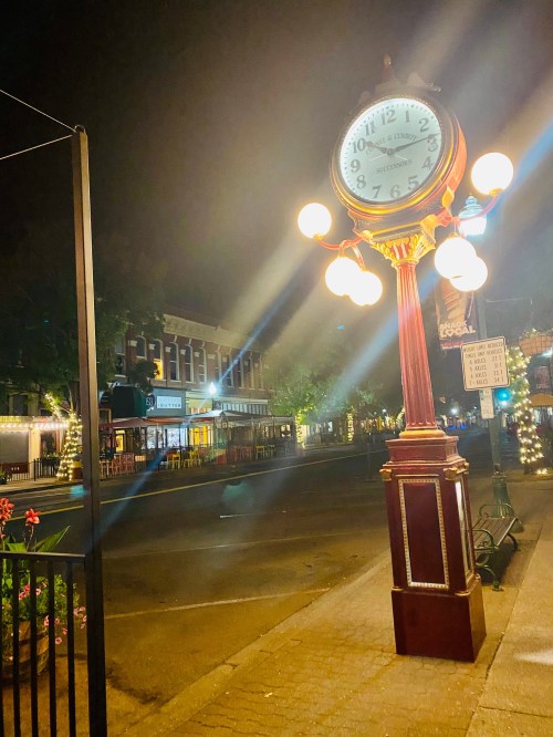 A vintage clock stands on a quiet street at night, illuminated by streetlights and surrounded by charming buildings.
