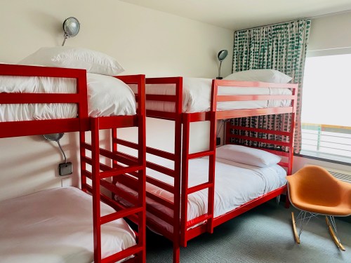 A brightly colored room with two red bunk beds and a modern orange chair, featuring white bedding and patterned curtains.