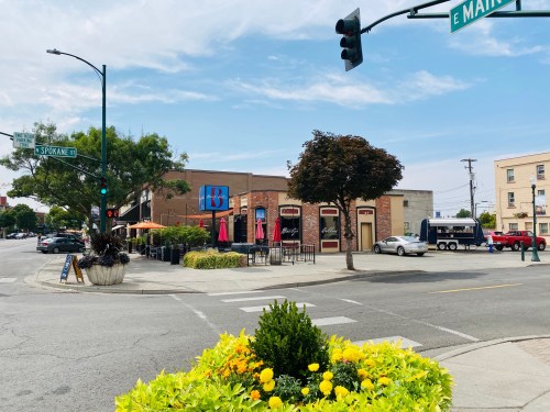 A street corner with a restaurant, outdoor seating, and colorful flowers in the foreground under a clear sky.