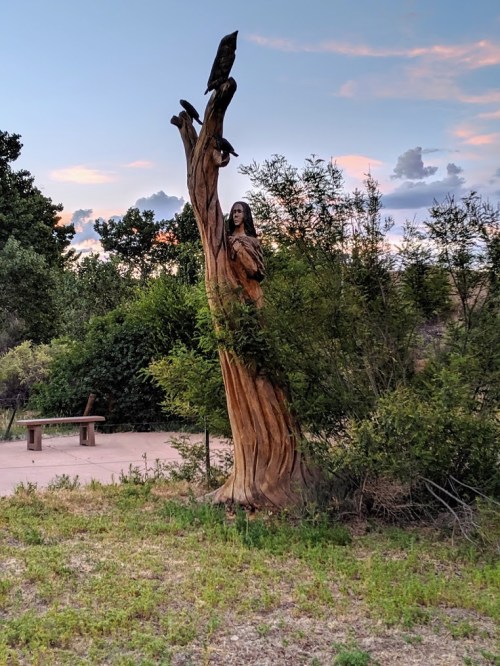 A wooden sculpture of a woman emerging from a tree, surrounded by greenery and a scenic sky.