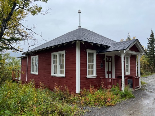 A small red wooden cabin with a gray roof, surrounded by greenery and trees on a cloudy day.
