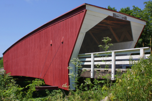 A red covered bridge with a white railing, surrounded by greenery under a clear blue sky.
