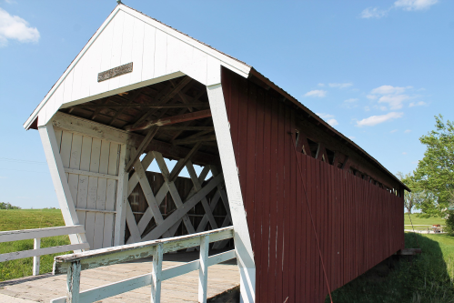 A red and white covered bridge with a wooden walkway, set against a clear blue sky and green fields.