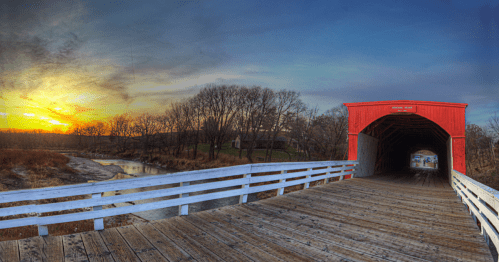 A scenic view of a red covered bridge at sunset, with trees and a stream in the background.