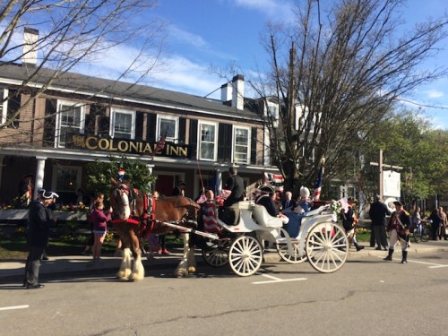 A horse-drawn carriage passes by the Colonia Inn, with people in historical attire celebrating a festive event.