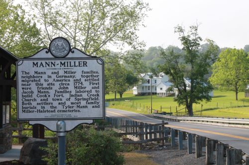 Historical marker for the Mann and Miller families, with a scenic view of a road and a white house in the background.
