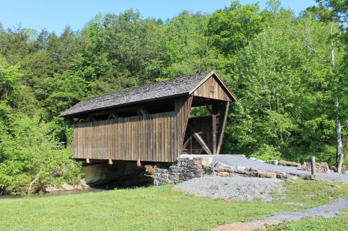 A rustic wooden covered bridge stands over a small stream, surrounded by lush green trees and a clear blue sky.