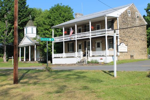 Historic stone building with a porch, American flags, and a nearby church, set in a green landscape.