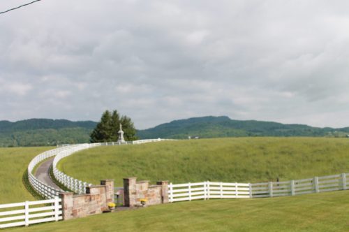 A winding white fence leads to a statue on a hill, surrounded by lush green fields and distant mountains under a cloudy sky.
