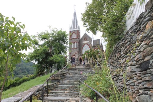 A stone pathway leads up to a historic church surrounded by greenery and trees on a cloudy day.