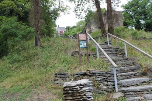 A stone pathway leads up a grassy hill, with a sign and a historic building visible in the background among trees.