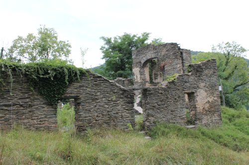 Ruins of a stone building overgrown with grass and vines, surrounded by trees and a cloudy sky.