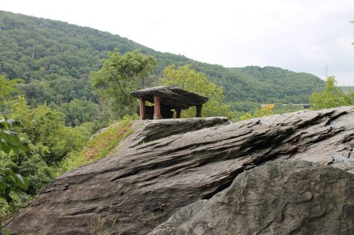 A rocky outcrop with a flat stone structure on top, surrounded by lush green hills under a cloudy sky.