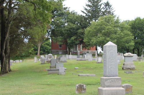 A peaceful cemetery with gravestones, trees, and a historic building in the background under a cloudy sky.