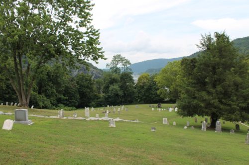 A serene cemetery with gravestones on a grassy hillside, surrounded by trees and overlooking a river and mountains.
