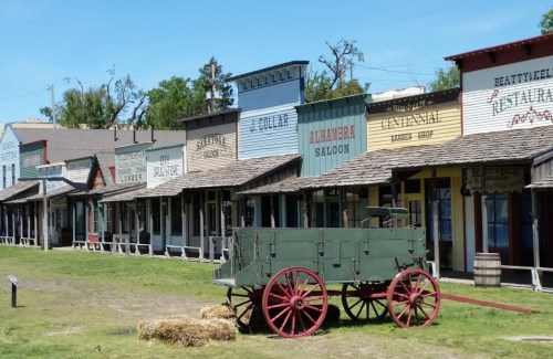 A historic western town with colorful storefronts and a green wagon in front, set on grassy land.