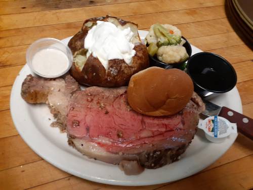 A plate featuring a large slice of prime rib, baked potato with sour cream, vegetables, a roll, and dipping sauces.