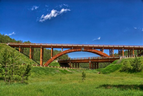 A wooden bridge arches over a green landscape under a clear blue sky, surrounded by trees and grass.