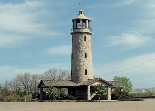 A tall stone lighthouse with a circular top, surrounded by trees and a clear blue sky.