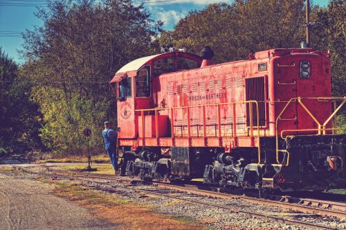 A vintage red locomotive parked on tracks, with a person in blue overalls inspecting it amidst autumn foliage.