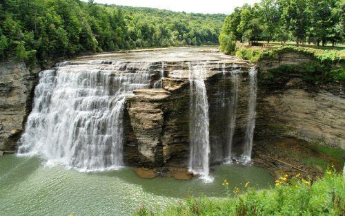 A scenic waterfall cascading over rocky cliffs into a serene green river, surrounded by lush trees and vegetation.