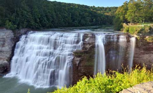 A scenic view of a waterfall cascading over rocky cliffs, surrounded by lush greenery and a clear blue sky.