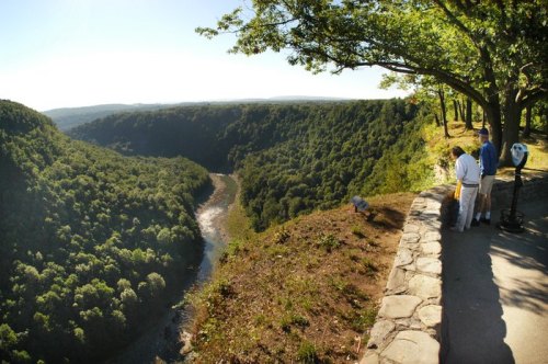 A scenic view of a river winding through lush green hills, with two people standing near a lookout point.