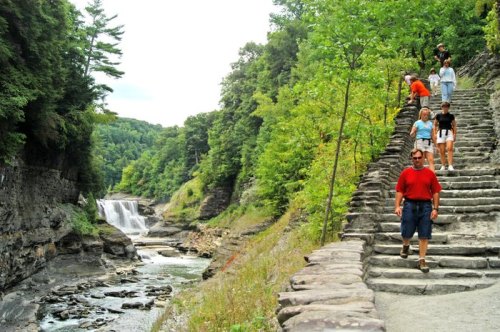 People walking down stone steps beside a river and waterfall, surrounded by lush green trees and rocky cliffs.