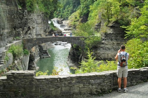 A person stands by a stone wall, taking photos of a scenic gorge with a bridge and waterfall in the background.