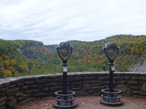 Two binoculars on a stone platform overlooking a colorful, forested valley under a cloudy sky.