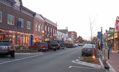 A charming street lined with shops and parked cars, illuminated by festive lights at dusk.