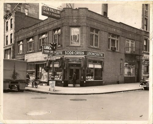 Black and white photo of a corner building featuring a soda fountain and luncheonette, with vintage signage and street activity.