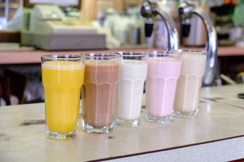 Five tall glasses of colorful milkshakes lined up on a counter, including yellow, brown, white, pink, and beige drinks.