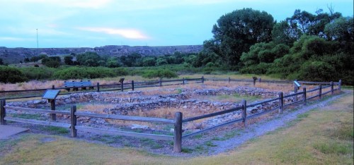 Ruins of a stone structure surrounded by a wooden fence, set in a grassy area with trees and hills in the background.