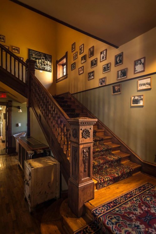 A wooden staircase with a patterned runner, surrounded by framed photos on the walls and warm yellow lighting.