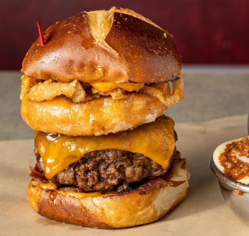 A towering burger with a beef patty, melted cheese, crispy onion rings, and a pretzel bun, served with a side of chili.