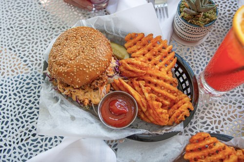 A sesame seed burger with coleslaw, pickles, and a side of sweet potato waffle fries and ketchup on a patterned table.