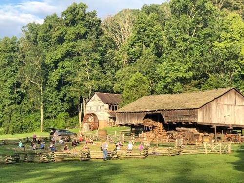 A group of people gathers near a rustic barn and watermill, surrounded by lush greenery and a clear blue sky.
